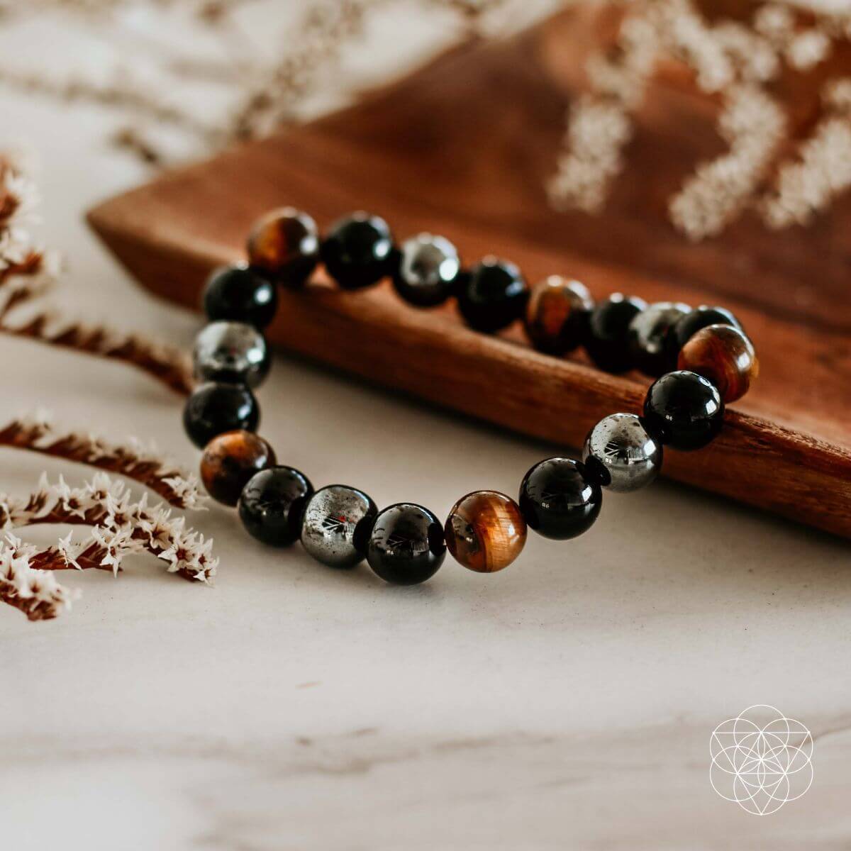 The Triple Protection Bracelet by Conscious Items, featuring alternating shiny black, brown, and metallic gray beads, is displayed on a wooden tray with dried white flowers on marble. The Conscious Items geometric logo sits in the bottom right corner.