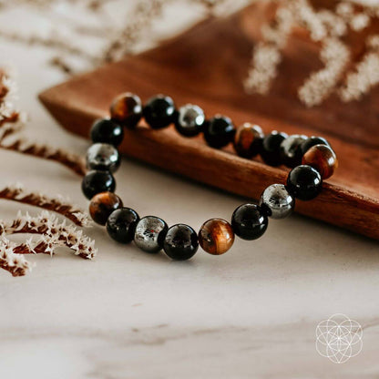 The Triple Protection Bracelet by Conscious Items, featuring alternating shiny black, brown, and metallic gray beads, is displayed on a wooden tray with dried white flowers on marble. The Conscious Items geometric logo sits in the bottom right corner.