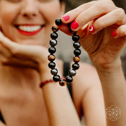 A smiling person with bright red lipstick and painted nails holds up a Conscious Items Peace & Protection Bracelet Collection, made of shiny black and brown stones, with their face softly blurred in the background.