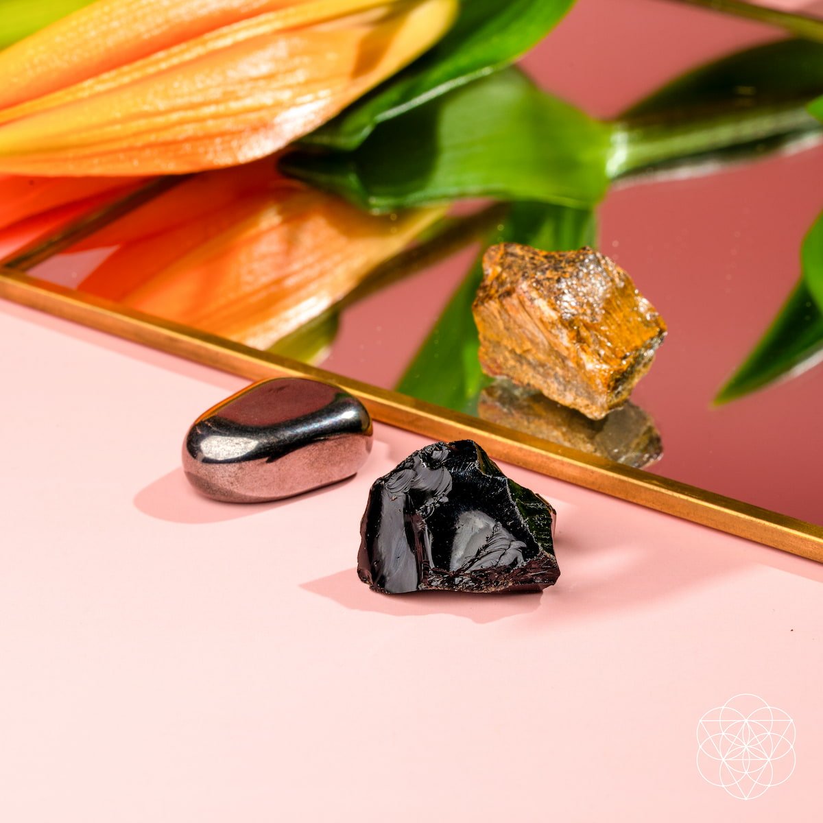 The Triple Protection Trio by Conscious Items—three unique stones—rests on a pink surface beside a gold-edged mirror that reflects an orange flower and green leaves.