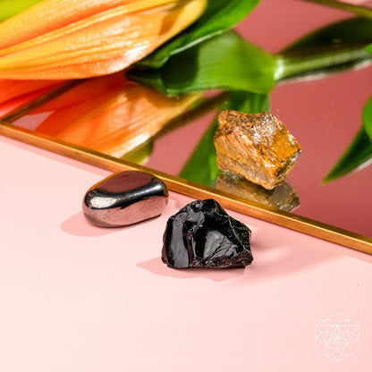 The Triple Protection Trio by Conscious Items—three unique stones—rests on a pink surface beside a gold-edged mirror that reflects an orange flower and green leaves.