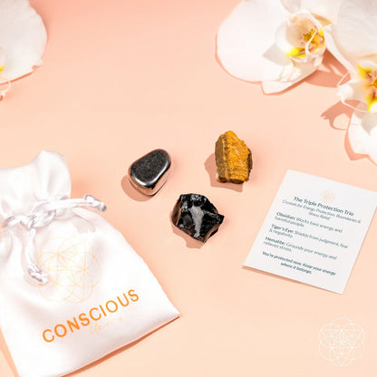 A white Conscious Items pouch sits beside The Triple Protection Trio stones, an info card, and white orchids on a peach background, showcasing energetic protection and elegance.