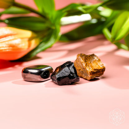 The Triple Protection Trio by Conscious Items features three grounding stones—silver and smooth, black and jagged, golden-brown and rough—resting on pink, with a blurred orange lily and green leaves in the background.
