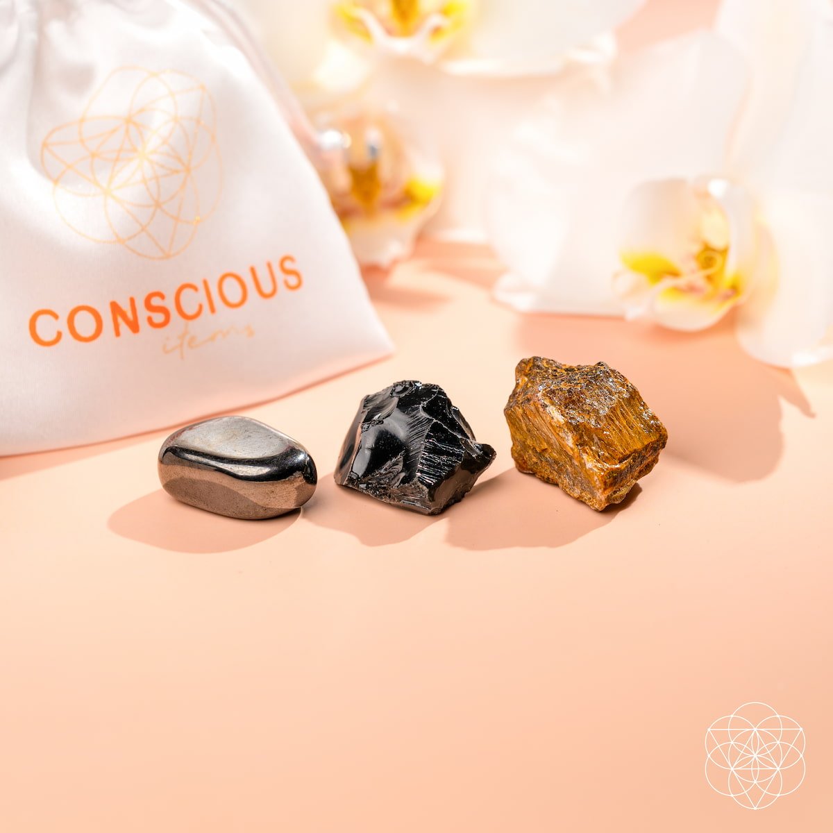 The Triple Protection Trio by Conscious Items—three polished grounding stones in metallic, black, and golden-brown—rests on a peach surface near a white CONSCIOUS ITEMS pouch, with white orchids in the background for energetic protection.