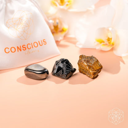 The Triple Protection Trio by Conscious Items—three polished grounding stones in metallic, black, and golden-brown—rests on a peach surface near a white CONSCIOUS ITEMS pouch, with white orchids in the background for energetic protection.
