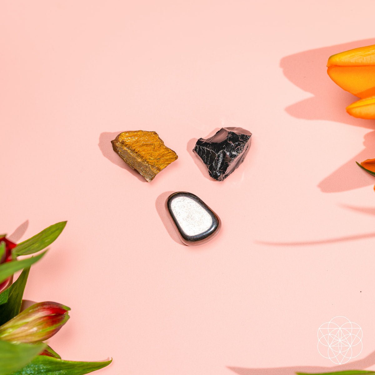 The Triple Protection Trio by Conscious Items features golden-brown, black, and white polished stones on a pink background with orange and red flowers in the corners, symbolizing energetic protection.