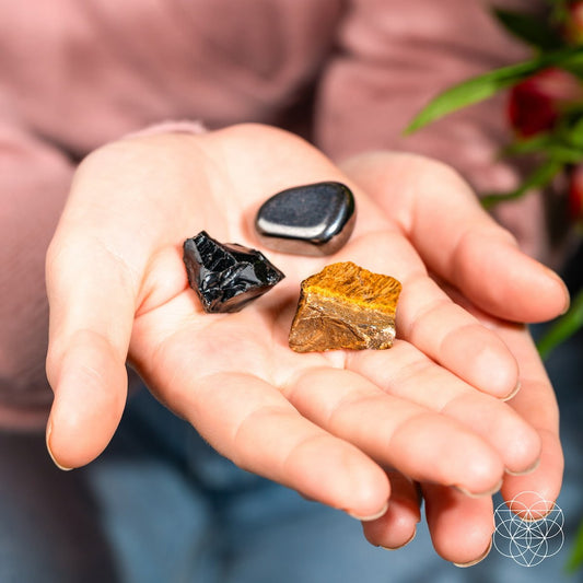 A hand displays The Triple Protection Trio by Conscious Items: three polished grounding stones—black, metallic silver, and golden-brown. In the softly blurred background are green leaves and a pink sleeve.