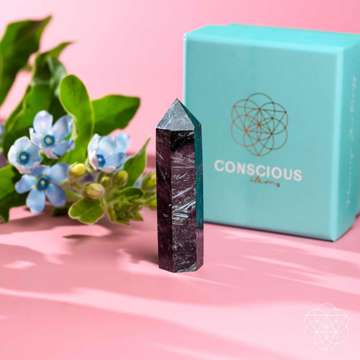 The Rare Arfvedsonite Point from Conscious Items stands upright on a pink surface, radiating breakthrough energy beside light blue flowers and a turquoise Conscious Items box with a gold geometric logo.