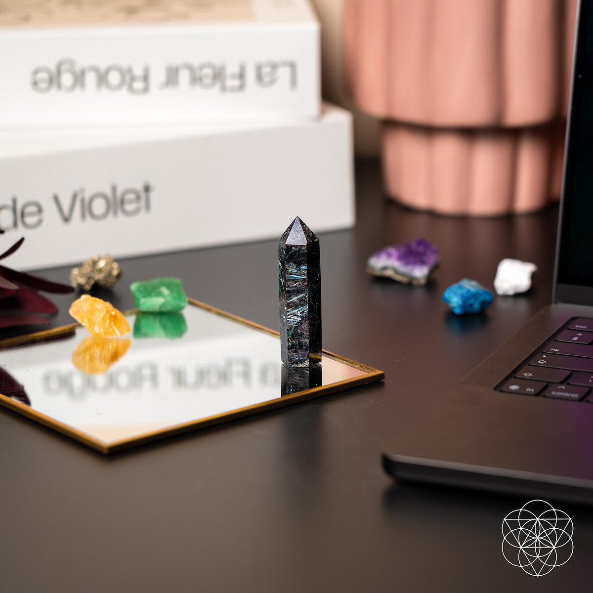The Conscious Items “This Is Your Breakthrough – Rare Arfvedsonite Point” stands on a mirrored tray beside colorful Compass Stones, with books, a laptop, and a copper vase—infusing your desk space with breakthrough energy.