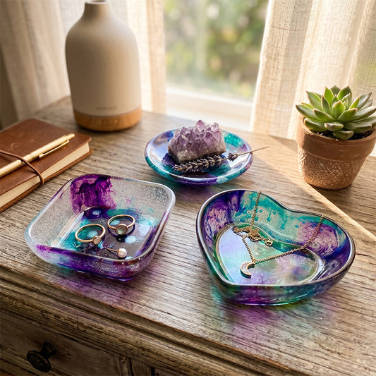 Three colorful jewelry dishes on a wooden surface with rings, necklaces, and crystals.