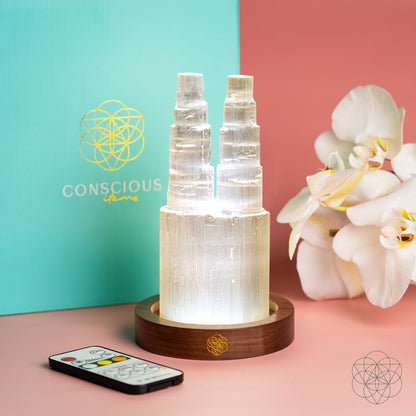 The Conscious Items Two-Tower Selenite Lamp – Deep Energy Cleanse & Reset stands on a wooden base beside a remote, white orchids, and a turquoise box with a gold logo—ideal for crystal cleansing.