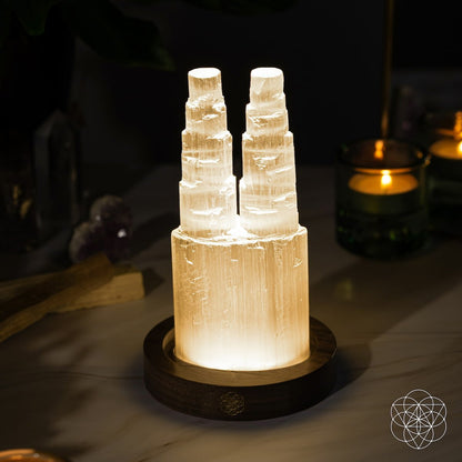 The Two-Tower Selenite Lamp – Deep Energy Cleanse & Reset by Conscious Items features twin crystal towers on a wooden base, ideal for crystal cleansing rituals and adding a soothing glow to your sacred space.