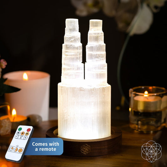 The Two-Tower Selenite Lamp – Deep Energy Cleanse & Reset by Conscious Items sits on a wooden base surrounded by candles, with a remote control included—ideal for energy cleansing rituals and crystal resets.