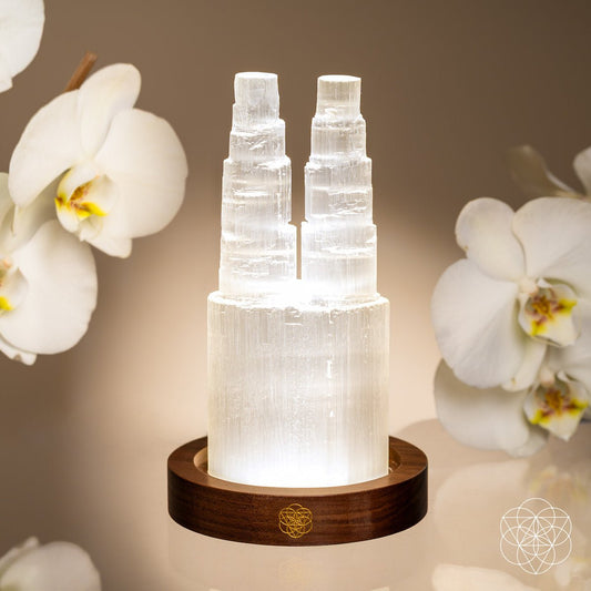 The Two-Tower Selenite Lamp by Conscious Items rests on a wooden base, accented with white orchids. Its gentle glow creates a tranquil mood, perfect for deep energy cleanse & reset in peaceful moments.