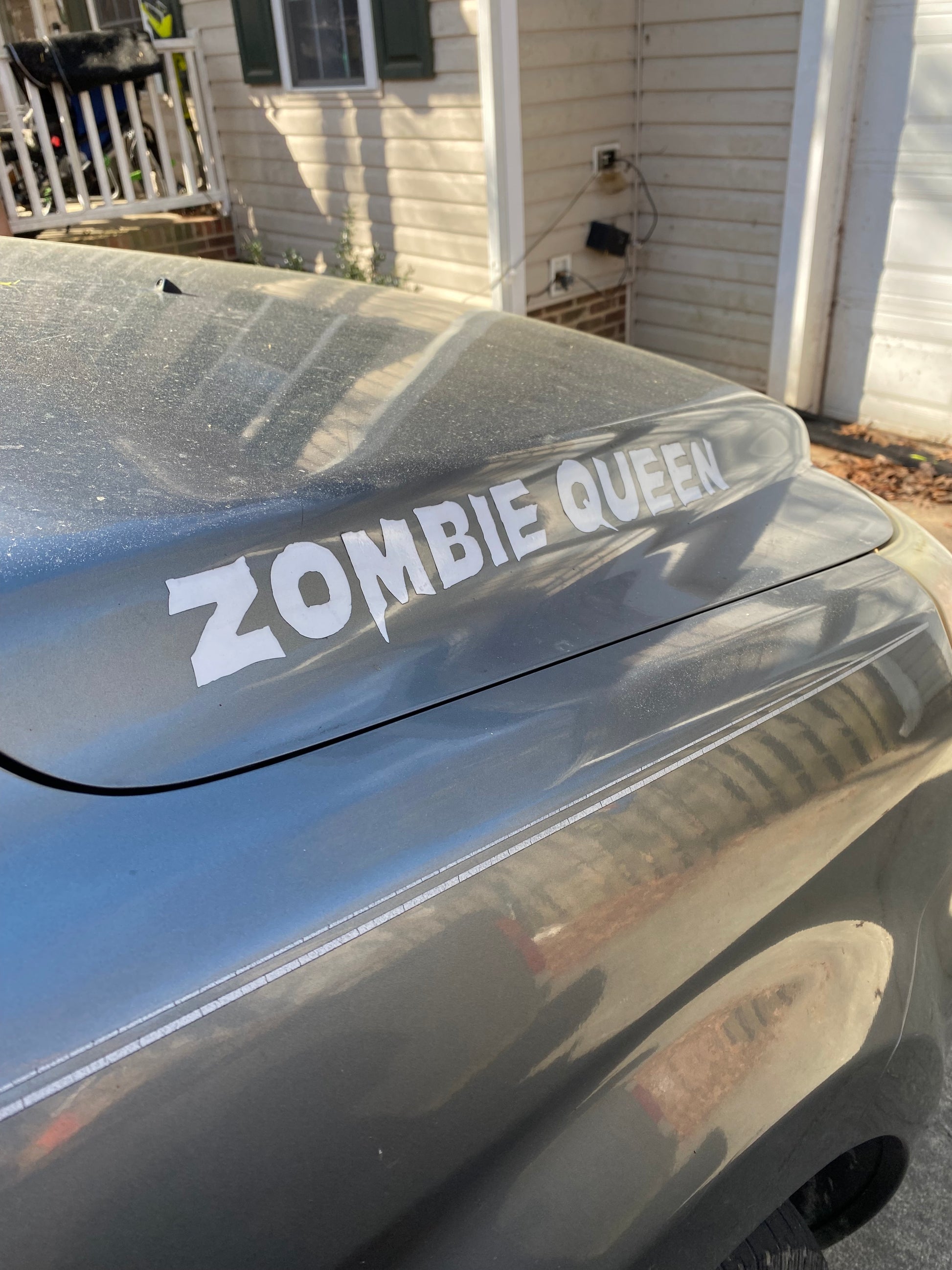A gray car featuring a “Zombie Queen” hood decal by GorgeousDragon Designs is parked in front of a house with beige siding and green shutters.